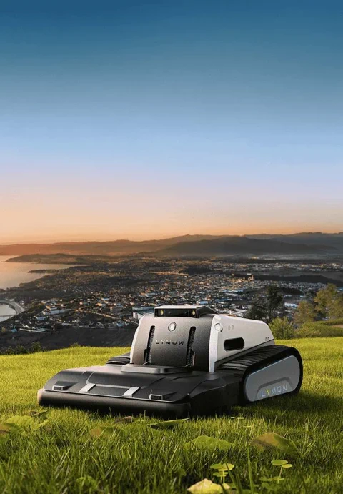 The Lymow One Plus robot mower, with its distinctive tank treads, sits on a hillside overlooking a city at sunset.